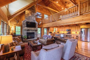 A spacious living area with a stone fireplace, a high ceiling with wooden beams, a dining area with a large table, and a kitchen with modern appliances.