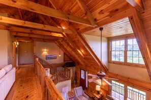 A spacious loft bedroom with a high ceiling, wooden beams, a large window, and a cozy seating area with a lamp.