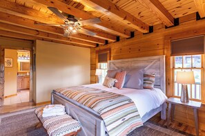 Une chambre à coucher dans une cabane en bois, comprenant un lit, une table de chevet, un banc, un ventilateur de plafond et une fenêtre avec des stores.