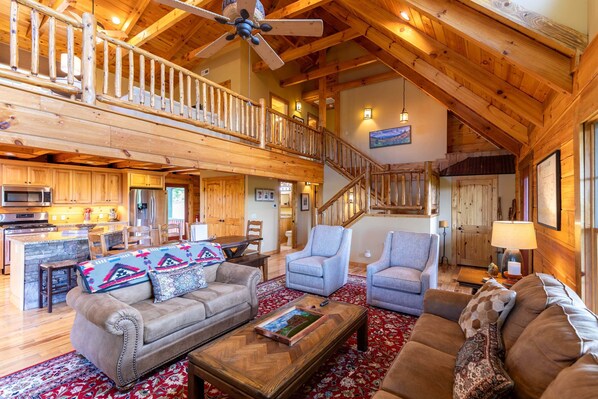 A spacious living area with a high ceiling, wooden beams, a staircase, and a kitchen with modern appliances.