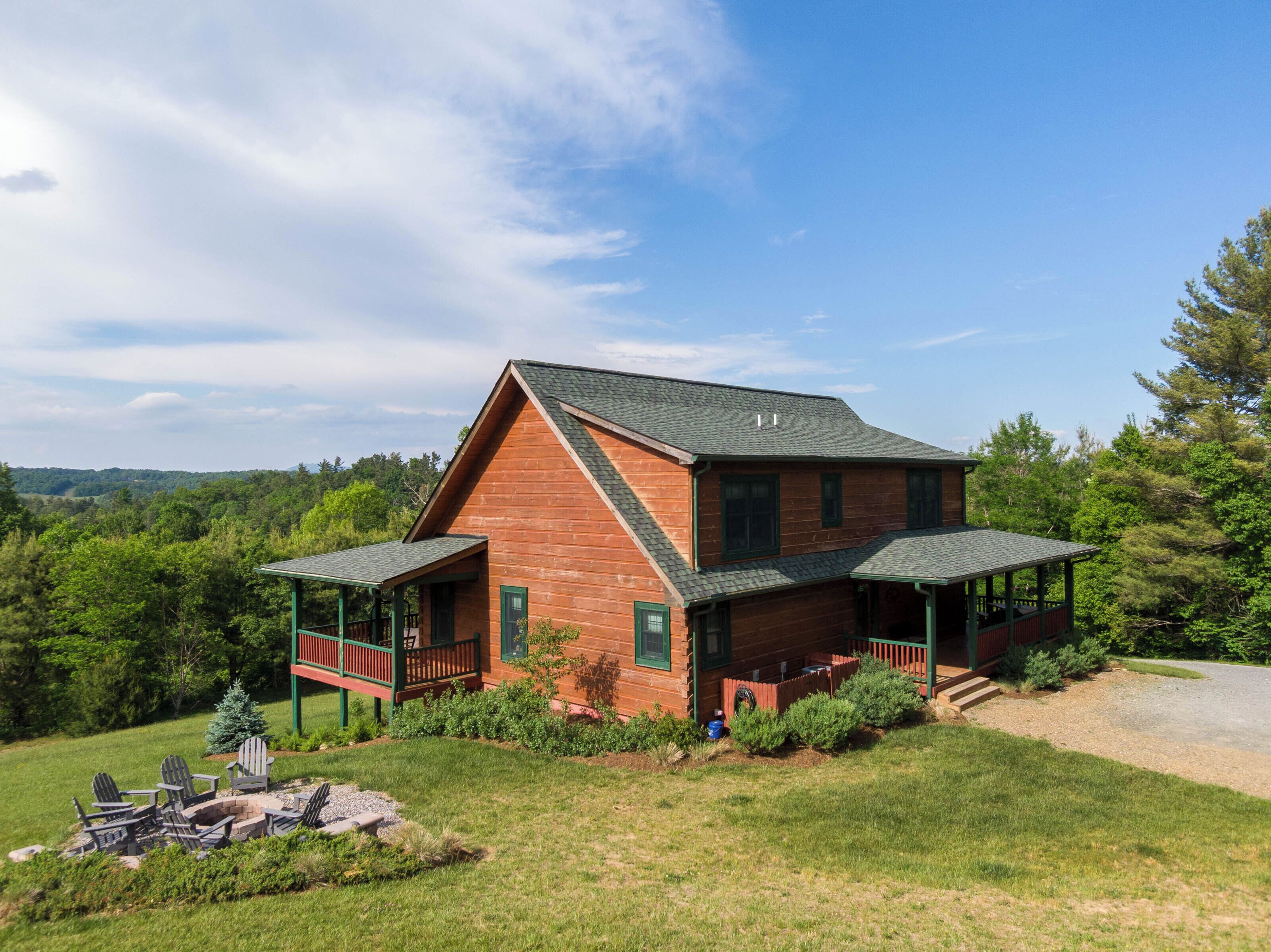 A two-story log cabin with a porch, a fire pit, and a view of the surrounding forest.