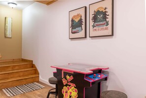 A room with a wooden staircase, a small table with arcade games, and two framed posters on the wall.