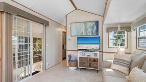 A modern living room with a sliding glass door leading to a balcony, a flat-screen TV mounted on the wall, a sofa with cushions, and a small side table.