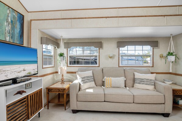 Una sala de estar con un sofá color beige, un televisor sobre un mueble blanco y dos ventanas con cortinas.
