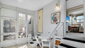 A bright, sunlit room with white walls and a large window, a staircase leading to an upper level, and a framed picture on the wall.