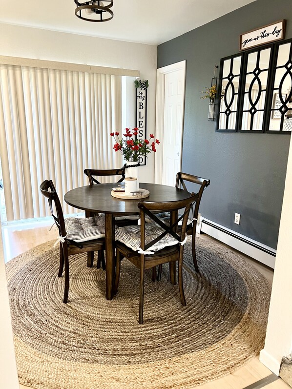 A dining area with a round table, four chairs, a woven rug, and a wall with a 'give us this day' sign. - Upper Grape, Midtown Home 2bed&1bath (Anchorage)