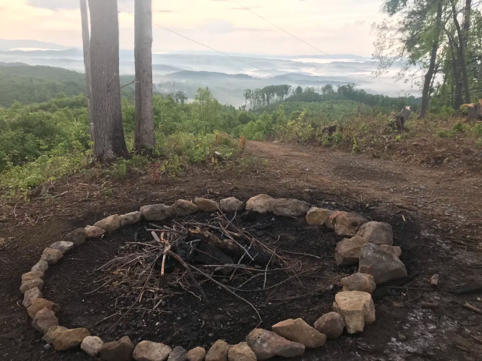 A circular stone fire pit surrounded by rocks, with a forested landscape and distant hills in the background.