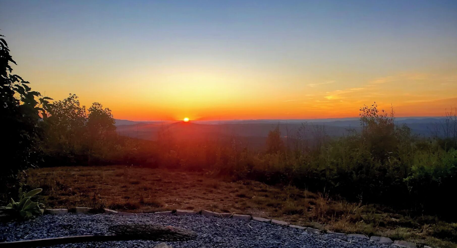 Un coucher de soleil sur un paysage vallonné, avec un chemin de gravier menant à une zone herbeuse.