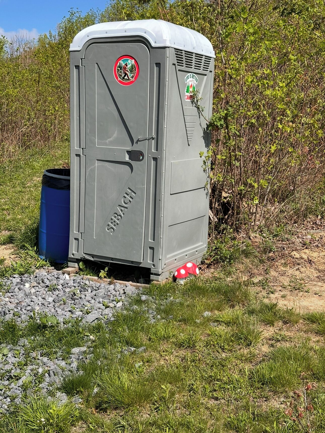 Un baño químico con un logotipo y texto en su lateral, ubicado al aire libre, rodeado de césped y arbustos.