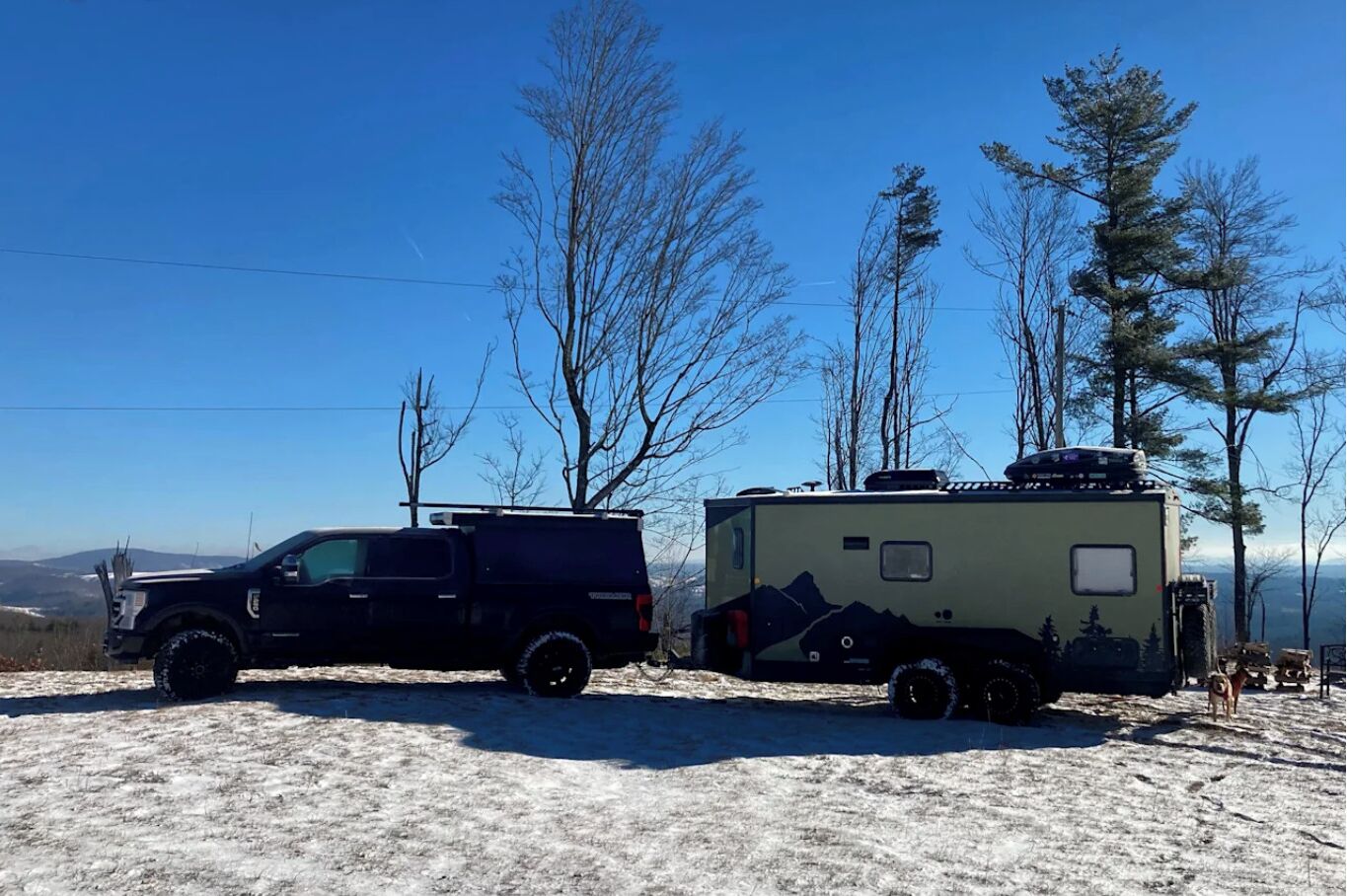 A black SUV and a green RV parked on a snowy field with trees and mountains in the background.