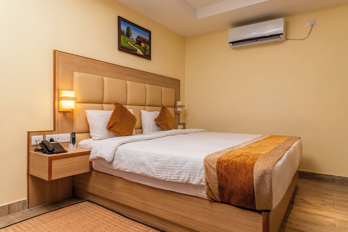 Une chambre d’hôtel avec une tête de lit en bois, du linge de lit blanc, un plaid marron, un téléphone sur la table de chevet et un appareil de climatisation.
