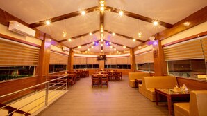 A spacious indoor area with wooden beams and a high ceiling, featuring multiple seating arrangements and a large windowed wall. - Lumbini Green Resort (Shivaraj)