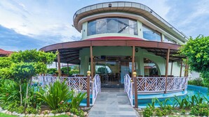 A two-story building with a curved roof and a glass facade, surrounded by a pool and lush greenery. - Lumbini Green Resort (Shivaraj)