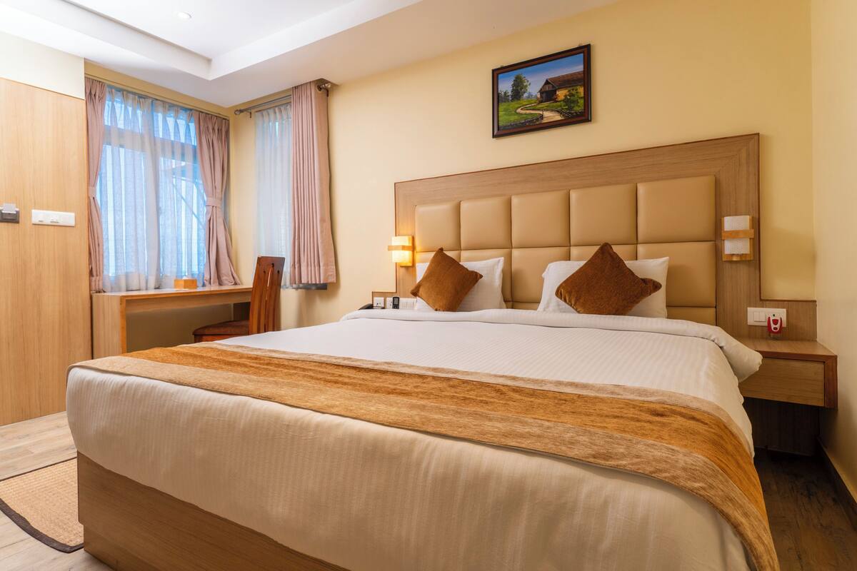 Une chambre d’hôtel avec un grand lit, une tête de lit en bois, deux tables de chevet avec des lampes, une fenêtre avec des rideaux et un tableau au mur.