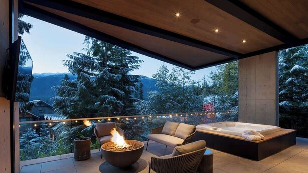 A modern outdoor living area with a fire pit, seating, and a hot tub, overlooking a snowy landscape.