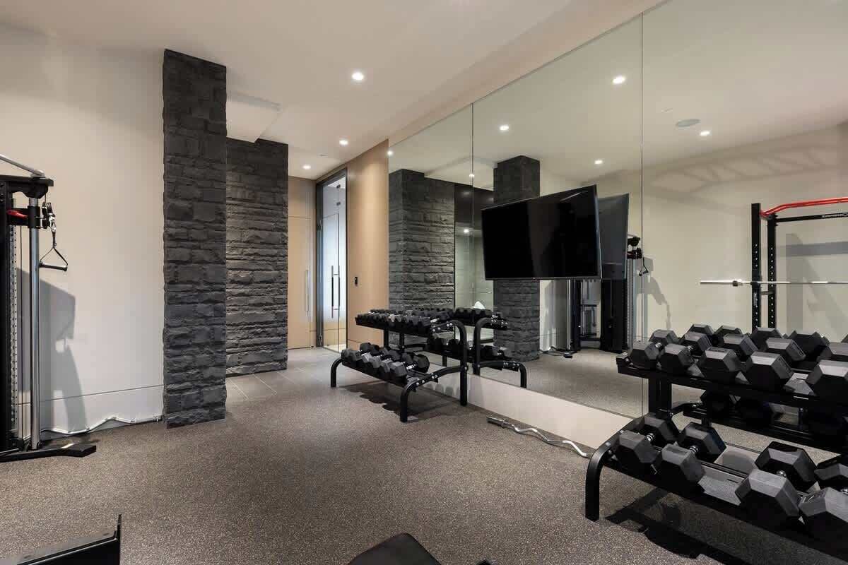 Ein moderner Fitnessraum mit Steinwänden, einem Fernseher und unterschiedlichen Trainingsgeräten.
