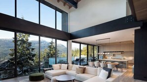 A modern living room with a sectional sofa, ottomans, and a dining area with chairs. Large windows offer a view of the outdoors.