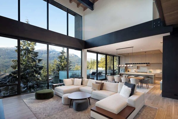 Un salon moderne avec un canapé d’angle, des poufs et un coin repas avec des chaises. De grandes fenêtres offrent une vue sur l’extérieur.