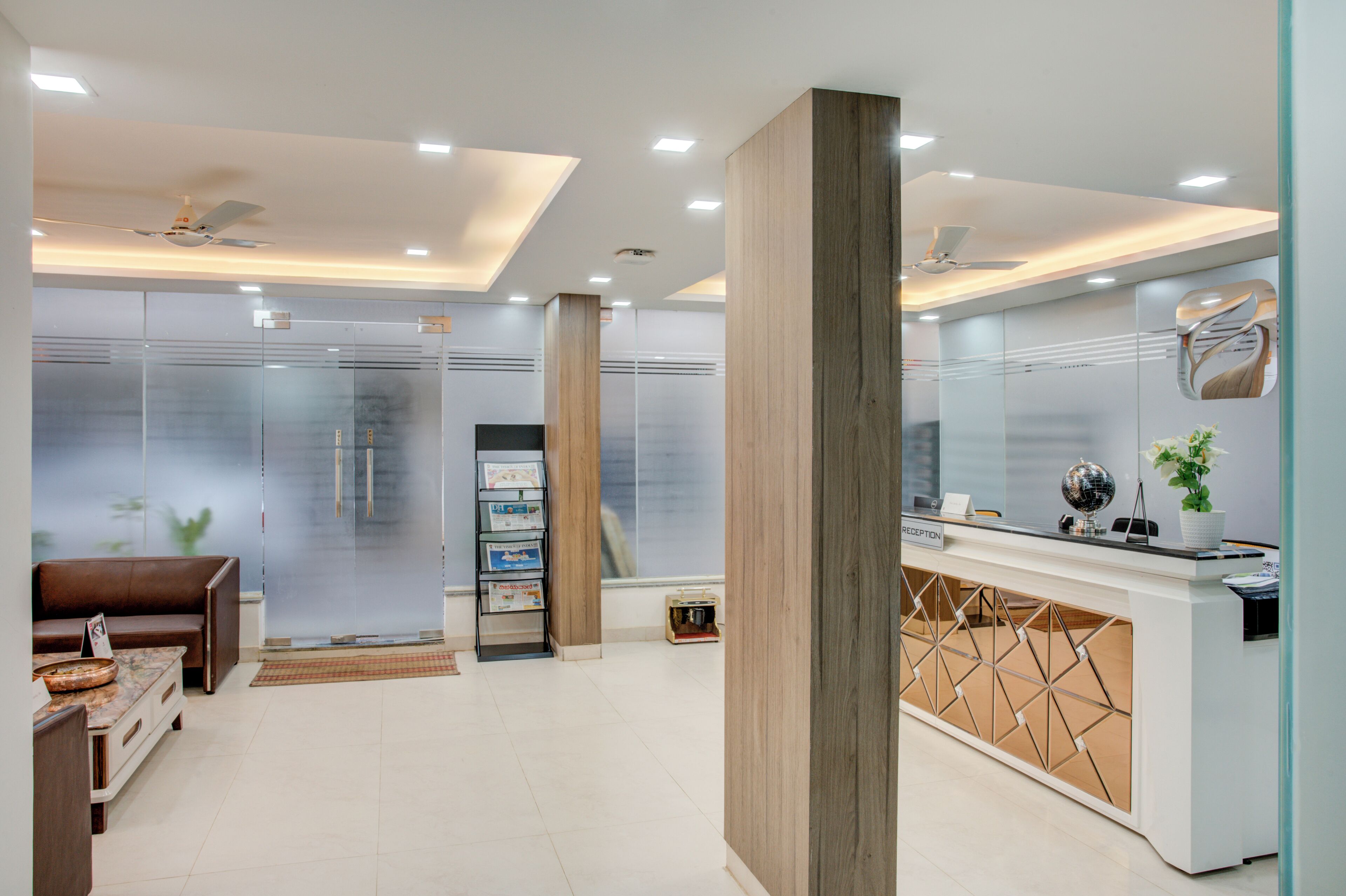 A modern reception area with a wooden reception desk, a glass partition, a seating area with a brown sofa, and a ceiling fan.