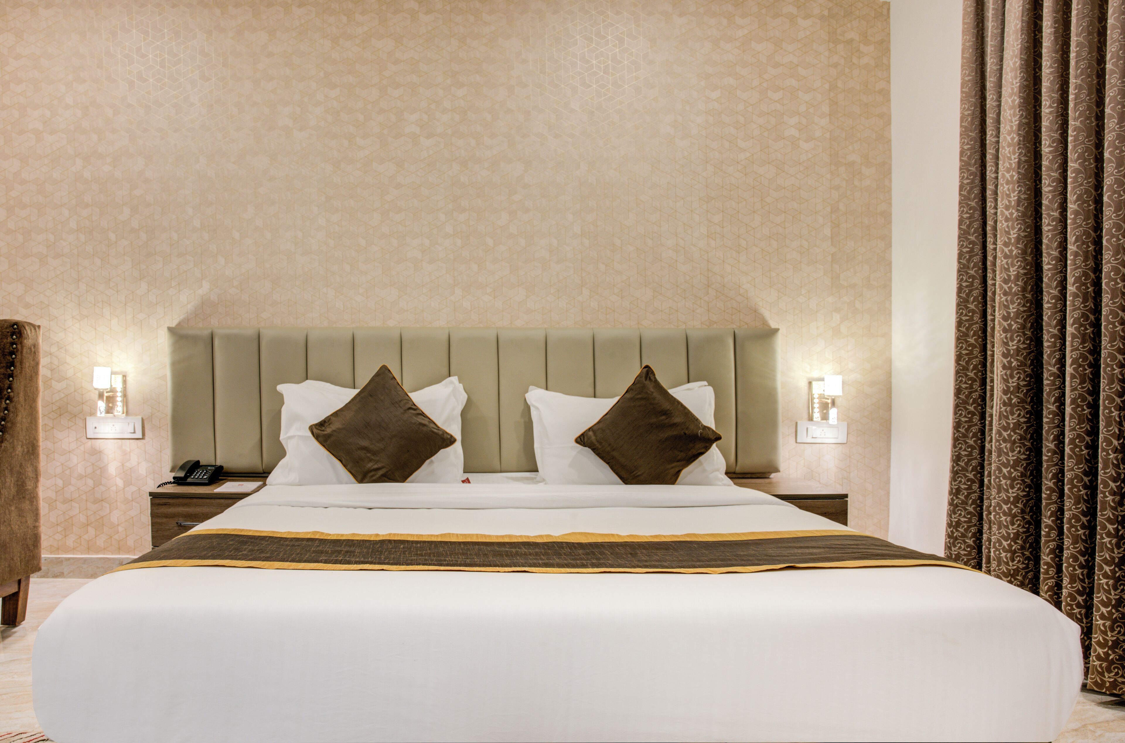 Una cama grande con sábanas blancas y dos almohadas marrones, una mesita de noche con un teléfono, y un aplique en la pared.