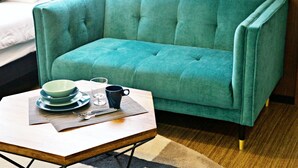 Een smaragdgroene bank met een houten salontafel ervoor, een bed met witte beddengoed, en een klein tafeltje met een kom, mok en bestek.