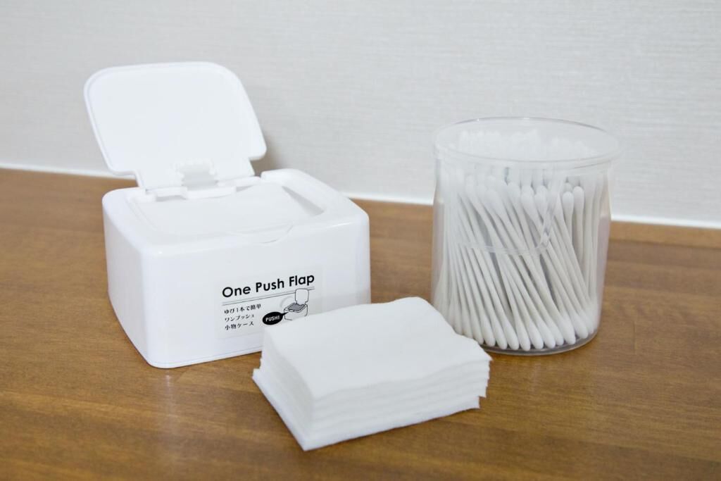 A white One Push Flap dispenser, a glass container with cotton swabs, and a stack of cotton swabs on a wooden surface.
