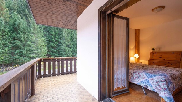 Een slaapkamer met een houten bed, nachtkastje, dressoir en een raam met uitzicht op een bos.