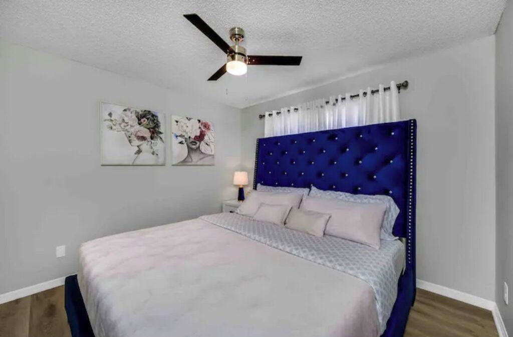 A bedroom with a tufted headboard, a bed with a patterned bedspread, a nightstand with a lamp, and two floral wall art pieces.