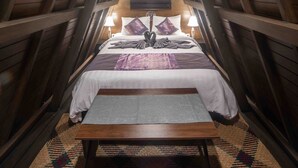 Un dormitorio en el ático con techo de madera, una cama con colcha blanca y morada, una mesita y una alfombra estampada.