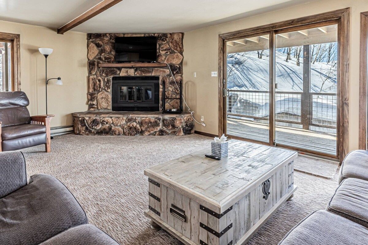 A living room with a stone fireplace, leather sofa, and a view of a snowy landscape through large glass doors.