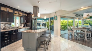 A modern kitchen with dark cabinets, a central island, and a dining area with a view of the outdoors. - Poseidon's Paradise: Luxury Waterfront Rental in Key West, Stunning Backyard Oasis and Private Pool! (Key West)