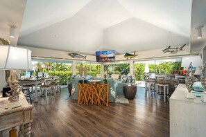 A spacious living area with a large window overlooking a tropical garden, a bar area with stools, and a dining table with chairs.