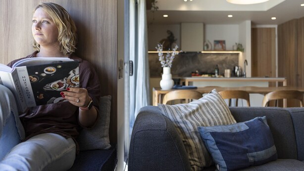 Una persona leyendo un libro en una sala de estar moderna con un sofá, un comedor y una cocina al fondo.
