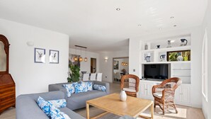 Een moderne woonkamer met een grijze bank, een houten salontafel, rieten stoelen en een keuken met witte kasten en decoratieve objecten.