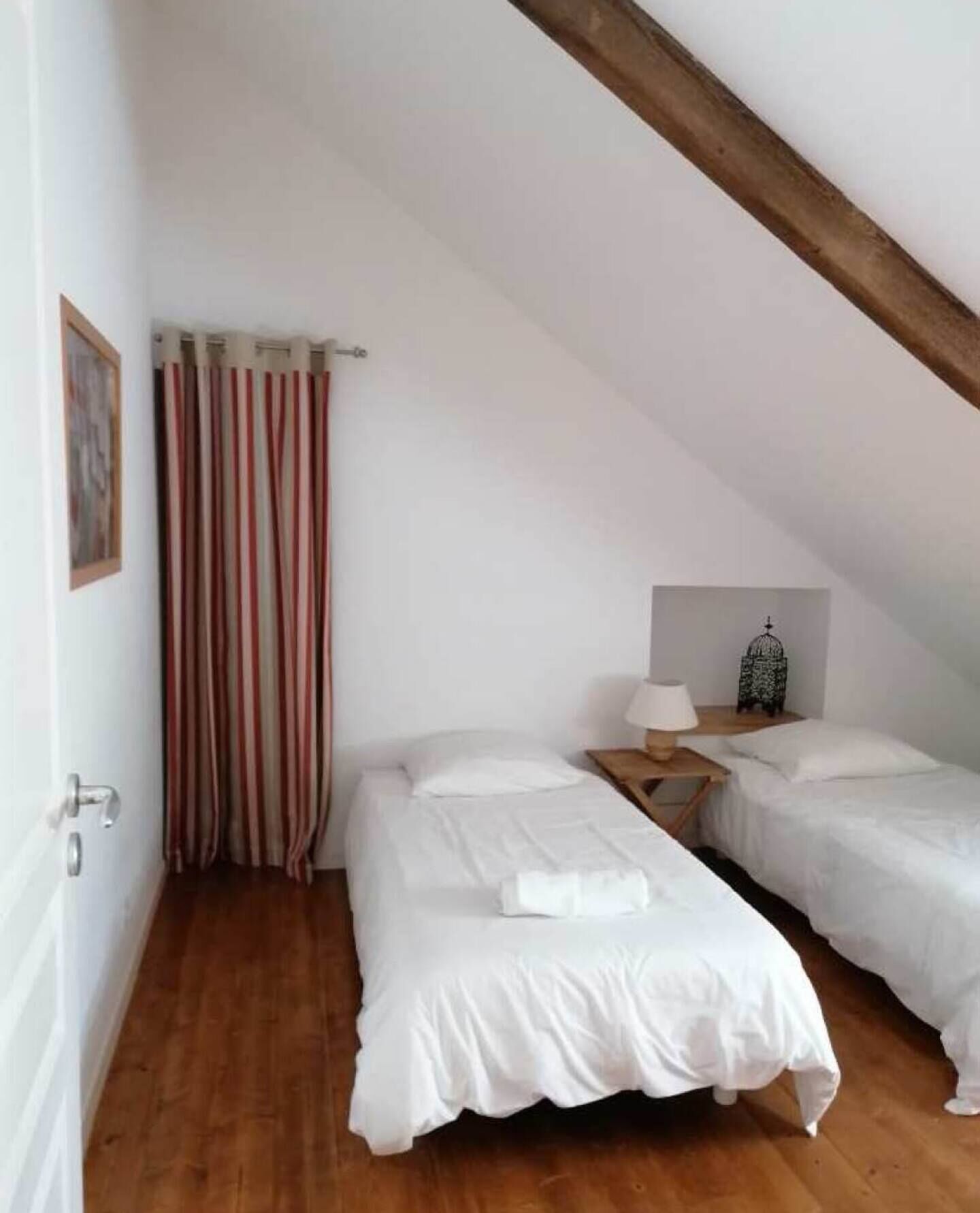 Een knusse kamer met twee eenpersoonsbedden, een houten vloer, een gestreept gordijn, een klein tafeltje met een lamp en een decoratieve vogelkooi.