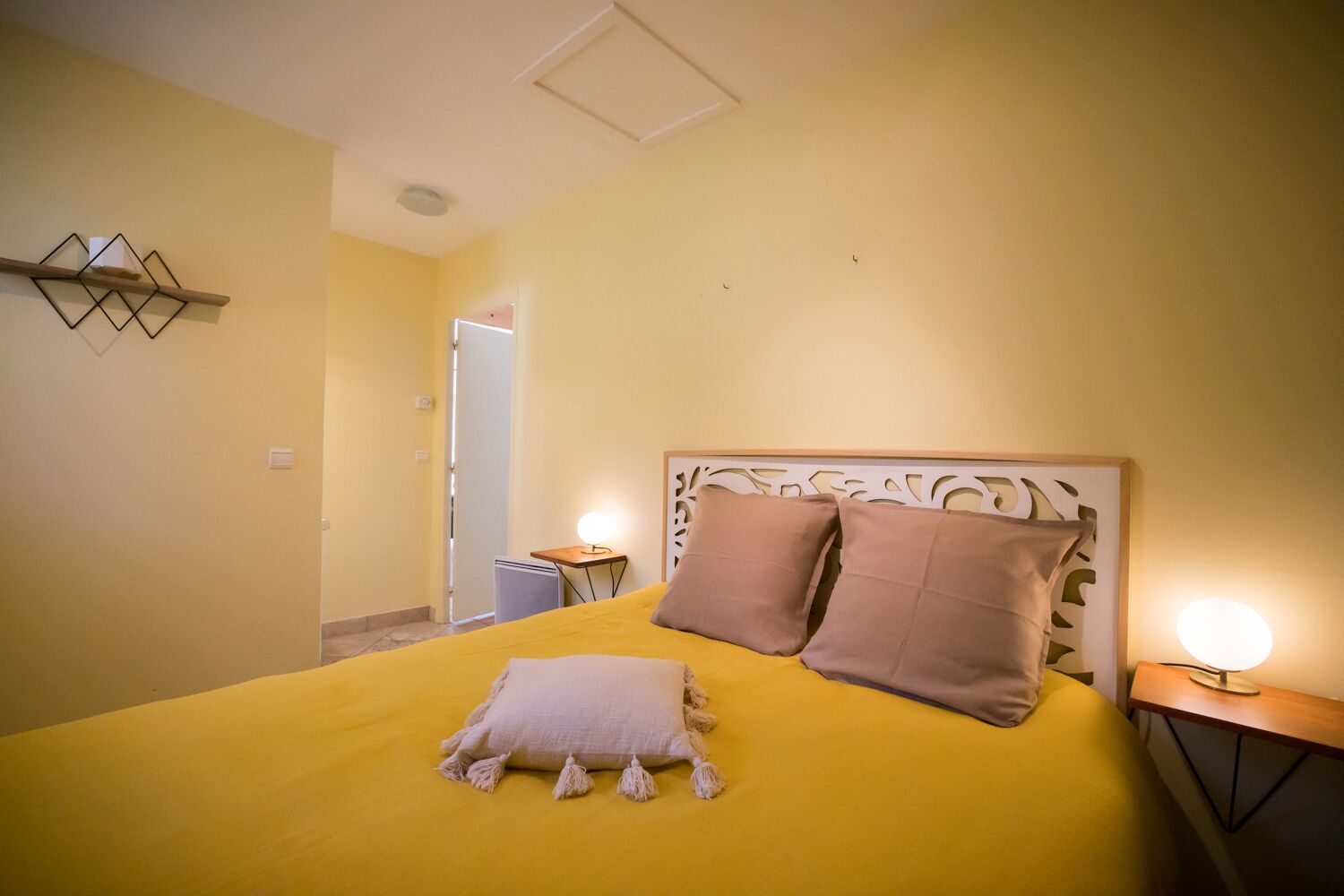 Een slaapkamer met een bed, twee nachtkastjes met lampen, een wandplank en een deur naar een andere kamer.