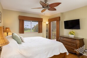 Un dormitorio con una cama grande, ventilador de techo, televisión, cómoda y ventana con persianas.