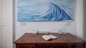 Un comedor con una mesa de madera, un libro, un florero de vidrio y un cuadro de una ola en la pared.
