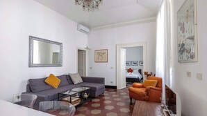 Un salón con un sofá gris, un sillón naranja y una mesa de centro. Hay un espejo grande en la pared, una araña y un suelo con un patrón hexagonal.