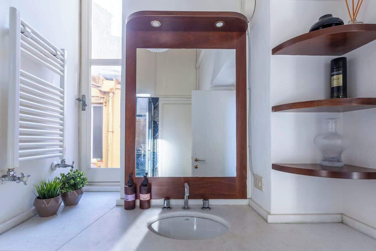 A bathroom with a large mirror, a white sink, two potted plants, and a wooden shelf with toiletries.