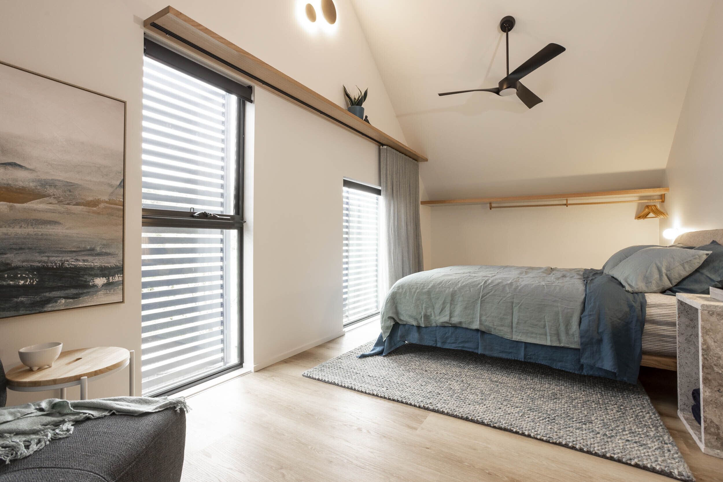 Ein modernes Schlafzimmer mit einem Bett, einem großen Fenster, einem Deckenventilator und einem Gemälde an der Wand.