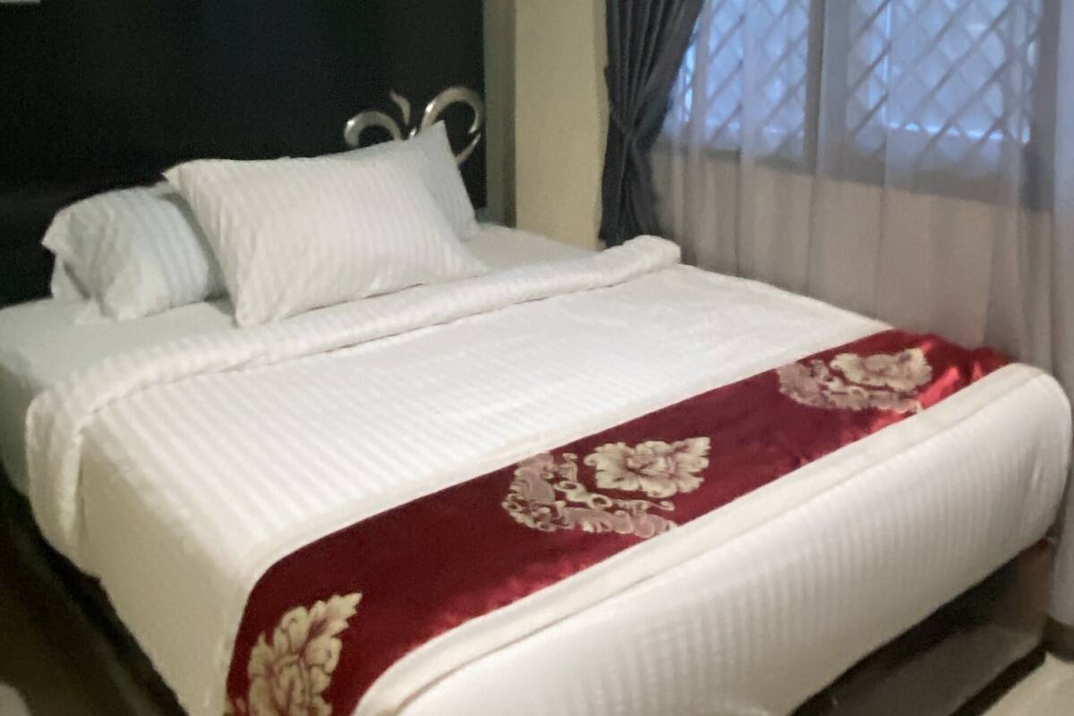 Una cama bien hecha con sábanas blancas y una colcha estampada de flores rojas, una pared verde y una ventana con mosquitero.