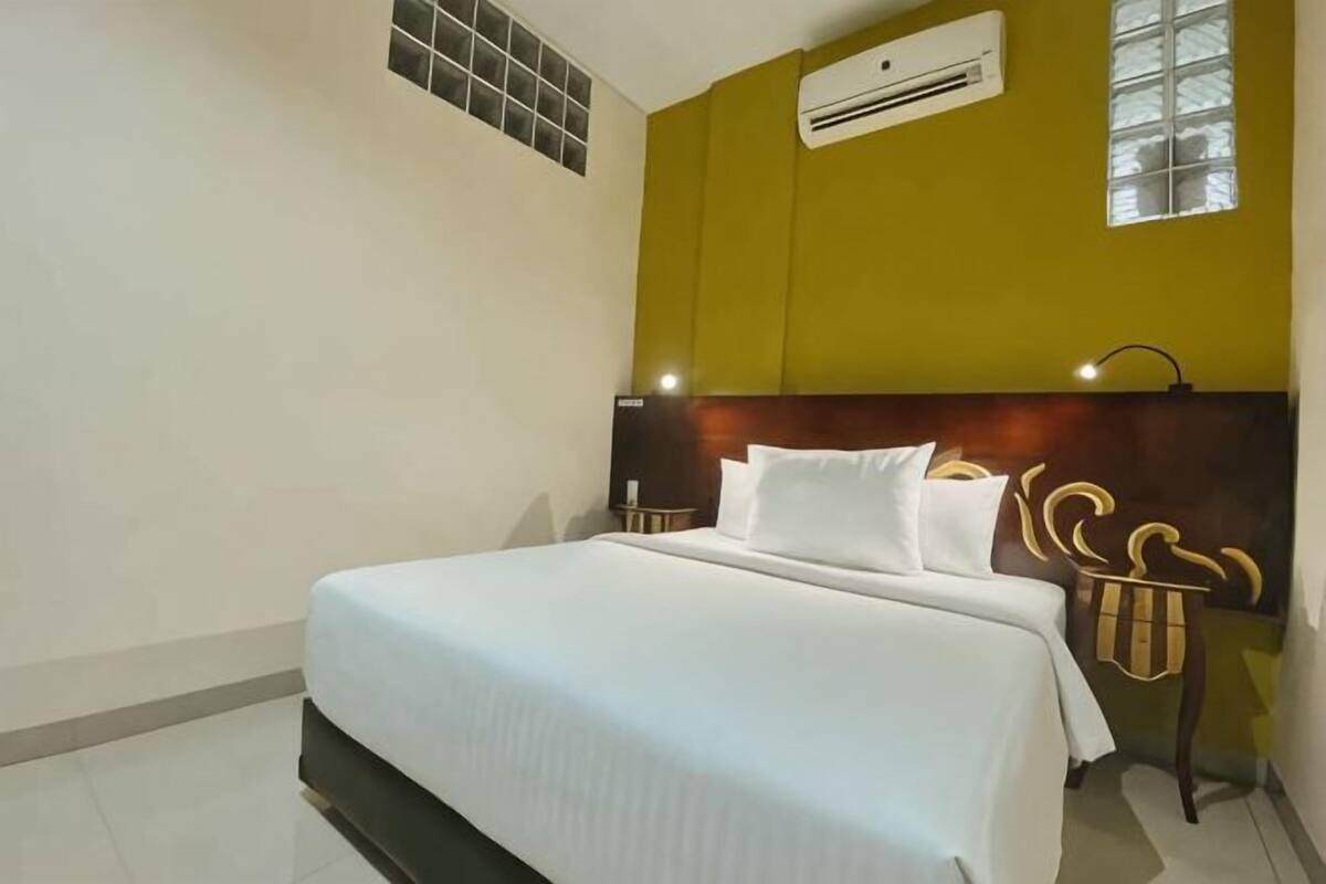 Habitación de hotel con cama, cabecero, unidad de aire acondicionado, ventana y lámpara de pared.