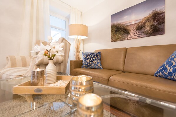 Ein Wohnzimmer mit einem beigefarbenen Sofa, einem Glastisch mit Dekoration, einer Lampe und einem Strandbild an der Wand.