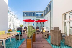 Eine Dachterrasse mit Korbmöbeln, roten Sonnenschirmen und Blick auf ein Gebäude im Hintergrund.