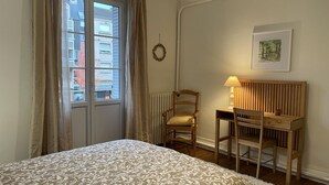 A bedroom with a bed, a desk, a chair, a lamp, a picture on the wall, and a window with curtains. - Apartment in Moulins: Comfort with Balcony, Linens and Towels Included (Moulins)