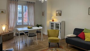 A living room with a sofa, a dining table with chairs, a chandelier, and a cabinet. - Apartment in Moulins: Comfort with Balcony, Linens and Towels Included (Moulins)