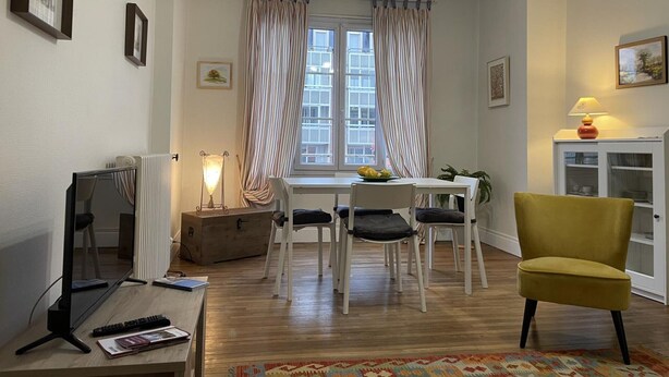 A dining area with a table and chairs, a window with curtains, a lamp, and a yellow chair. - Apartment in Moulins: Comfort with Balcony, Linens and Towels Included (Moulins)