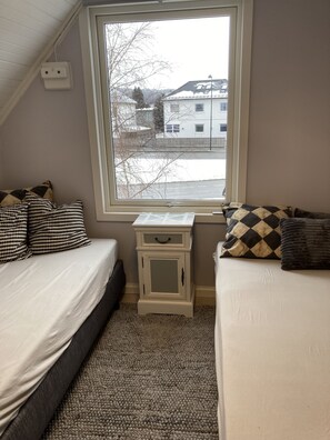 A bedroom with two beds, a window with a view of buildings, a small white nightstand, and a gray carpet.