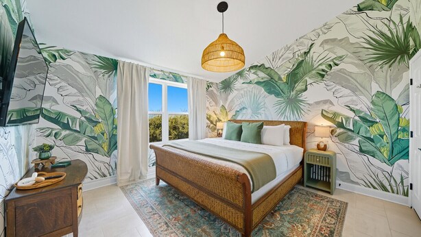 A bedroom with a large bed, a wooden nightstand, a wall-mounted TV, and a window with curtains. - Ciel - Endless Holiday Stays! (St. Augustine Beach)